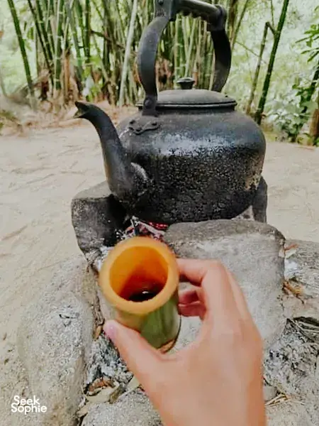 Close-up of a hand offering a small cup as a kettle pours over a campfire in Khao Lak during an outdoor stop
