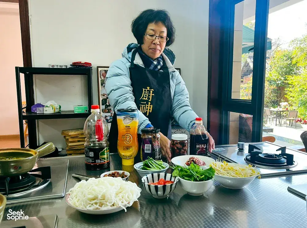 Fancy preparing a Yunnan meal in a bright Lijiang kitchen, arranging noodles and vegetables beside bottles and utensils