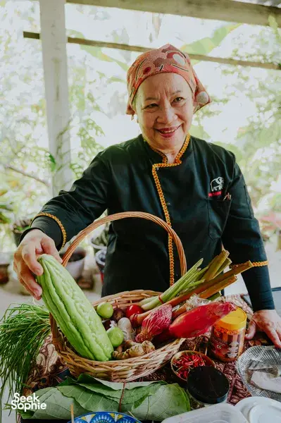 Seorang tukang masak pribumi Sabah di Kota Kinabalu memegang bakul berisi sayur-sayuran hijau dan herba segar, memakai tudung kepala serta pakaian tradisional