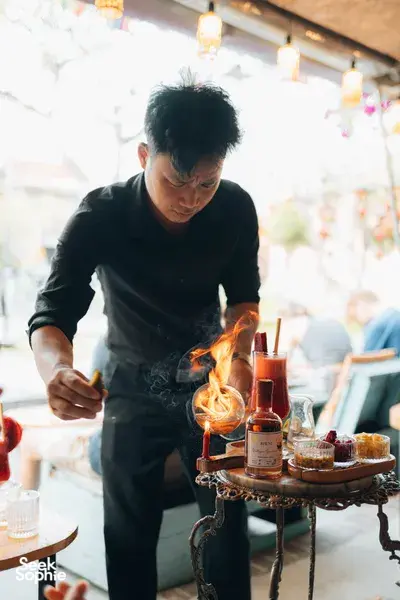 Man igniting a flaming cocktail in a glass beside bottles during a Hoi An cocktail tour at a hidden bar