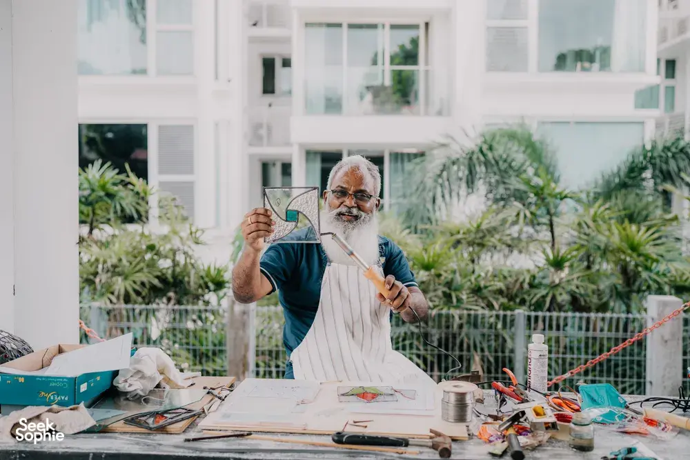 Elderly glass artist shaping a curved stained-glass panel at a table in a bright garden, Singapore—workshops in Singapore