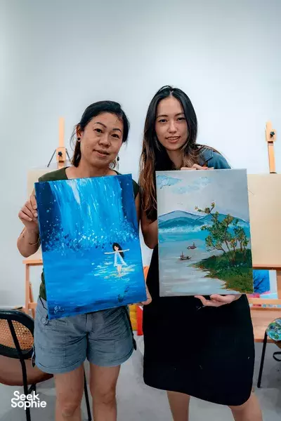 Smiling duo holding blue landscape canvases at CocoArt SG art studio in Singapore, after private art jamming workshops in Singapore