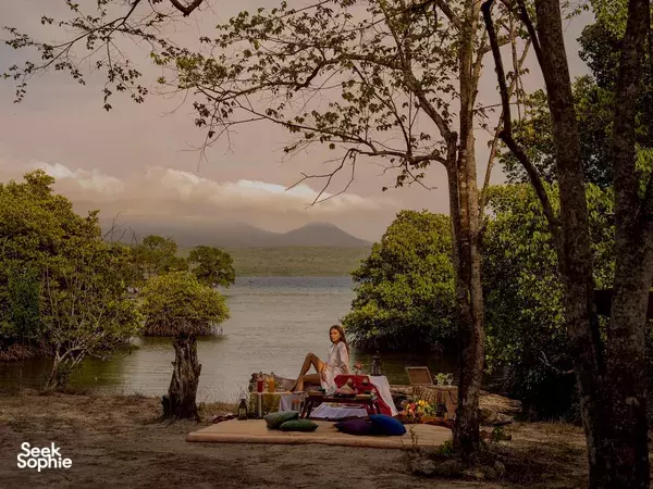 Tranquil lakeside retreat in Indonesia with a guest seated on a yoga mat by still water beneath tall trees at dusk