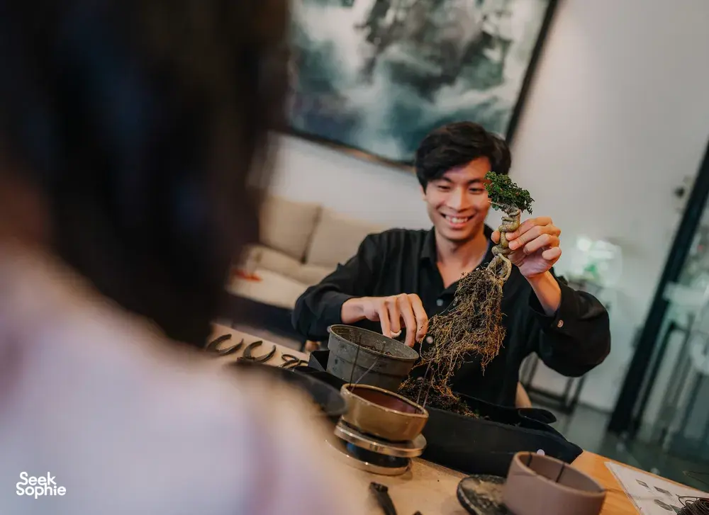 Smiling participant holding a tropical bonsai plant over a worktable of pots and tools during a Japanese-style workshop in Singapore