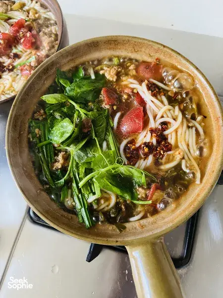Close-up of steaming Chinese noodle soup in a clay pot with greens, herbs and chilli oil, with another bowl nearby in China