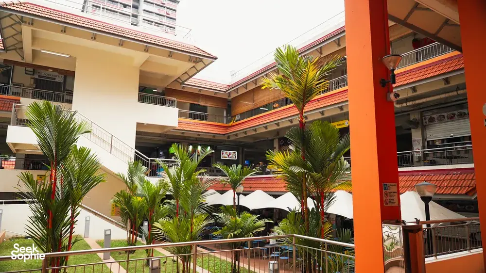 Open-air hawker centre courtyard in Singapore with palm trees, walkways and seating around an orange column — Singapore hidden gems