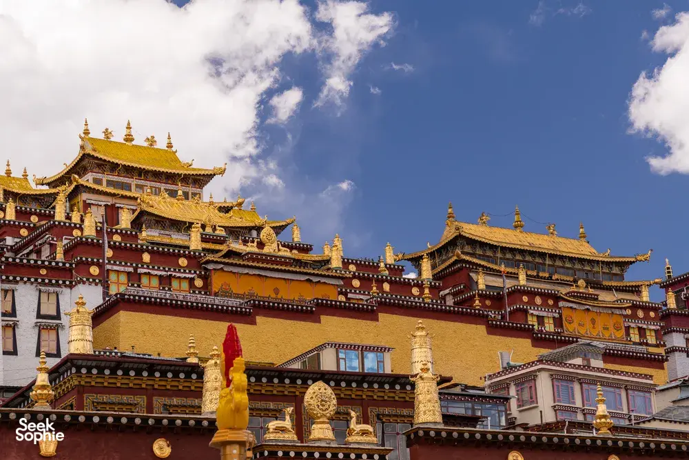 Ornate golden-roofed Tibetan-style palace buildings in Shangri-La against a bright blue sky, with scattered clouds overhead