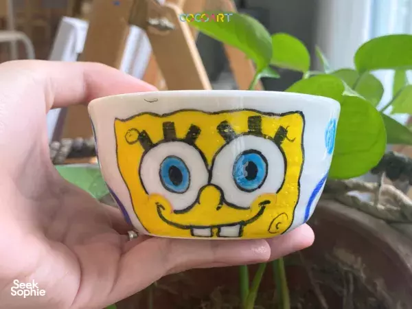 Hand holding a small glazed ceramic bowl painted with a smiling cartoon face, made in a Singapore pottery studio workshops in Singapore