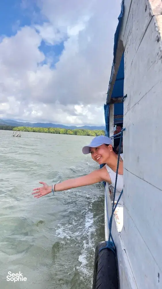 Seorang pengembara tersenyum membongkok di atas badan perahu layar putih di perairan hijau yang beralun semasa pelayaran di bawah langit biru, Labuan Bajo