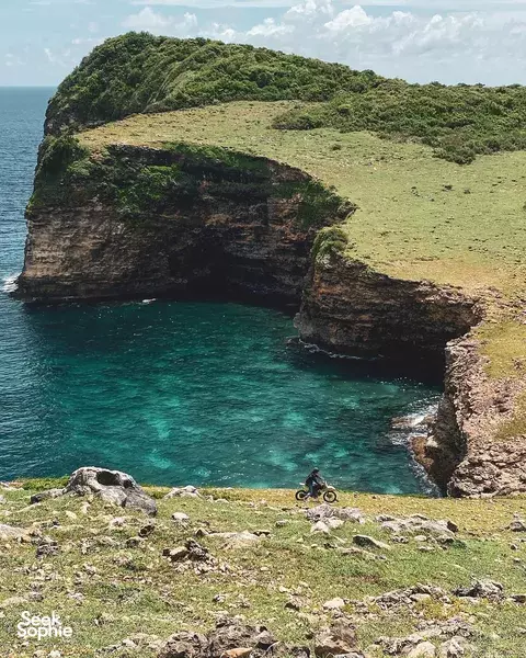 Motorcycle tour riders pause atop a cliff over turquoise bay on Lombok, with a sweeping coastline and sea below