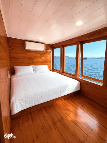 Wooden phinisi bedroom with double bed, bedside lights and sea-view windows on La Famille’s 3D2N shared Komodo cruise from Labuan Bajo