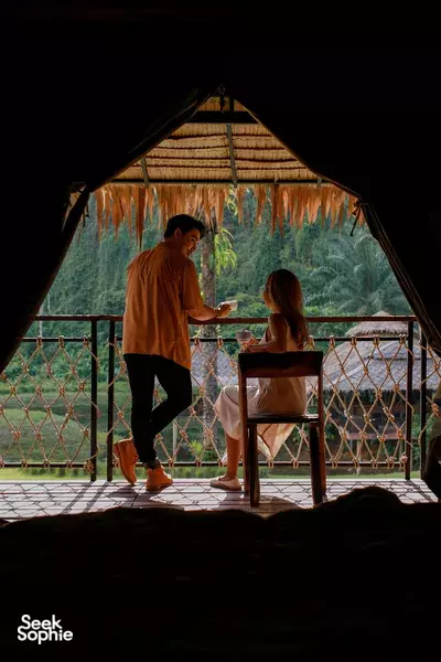Couple chatting on a wooden veranda table under hanging curtains, framed by trees and misty hills at dawn, in Khao Sok National Park