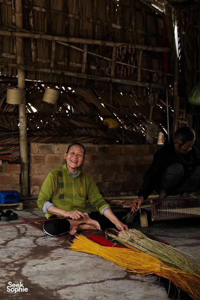 Traditional craft stall in Hoi An at dawn, with a smiling worker arranging yellow straw bundles in a rustic workshop