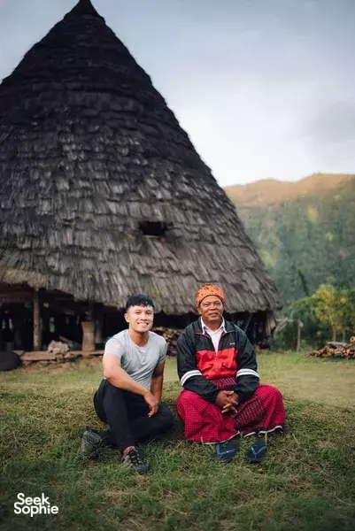 Two men kneeling and smiling outside a traditional thatched house, with green hills behind on a Labuan Bajo trip