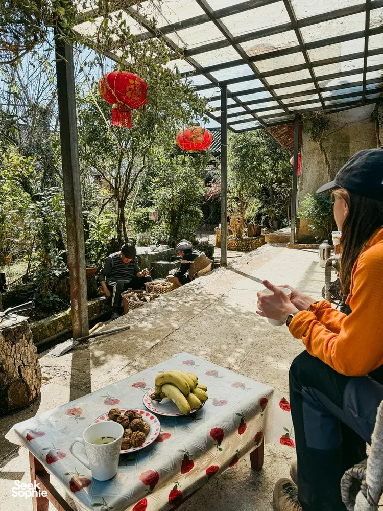 Orange-jacketed traveller reaching for food at a table in a shaded guesthouse courtyard in Lijiang during the village trek