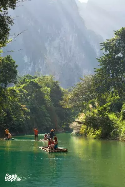 Longtail boats drifting across calm green water with towering misty limestone cliffs in the distance, at Cheow Lan Lake