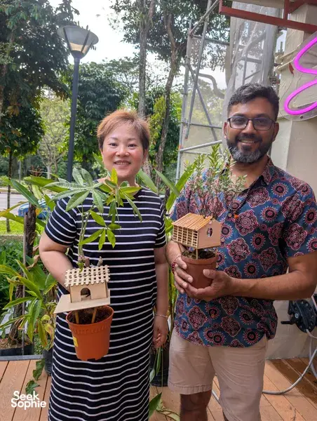 Smiling participants hold potted saplings beside a finished mini treehouse at a secret farm in Singapore, surrounded by tropical greenery