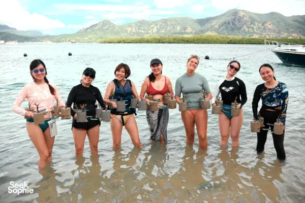 Travellers standing knee-deep in clear water holding planted coral during a reef-restoration day trip near Rinca, on Komodo Island