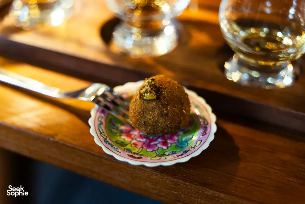 Decorative tasting plate with a small bite beside amber whisky glasses on a wooden bar, during a small-batch whisky tasting in Singapore