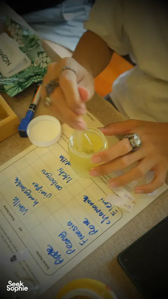 Hands stirring yellow wax in a glass jar over a recipe sheet for scented coco-soy candles, workshops in Singapore