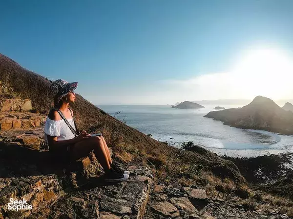 A traveller sitting on a rocky slope above a rugged coastline, watching waves roll in on Komodo Island