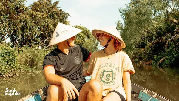 Two travellers in conical hats chatting on a covered boat during a Mekong Delta wildlife boat ride through greenery