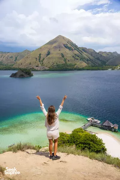 Woman overlooking a bright turquoise bay from a grassy cliff above a white-sand beach during a private Komodo cruise, Komodo Island