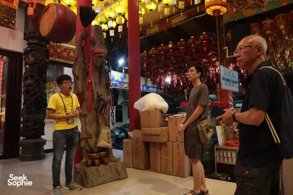 Hey Lad Tour guide and visitors gathered beside a large Chinese deity statue in a lantern-lit temple during night rituals in Singapore, Singapore hidden gems