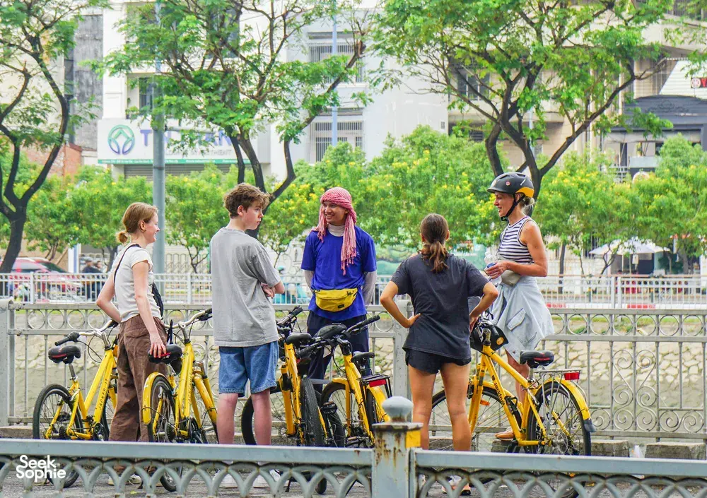 在胡志明市绿意盎然的河畔,一群骑行之旅的游客站在黄色自行车旁,与当地向导交谈