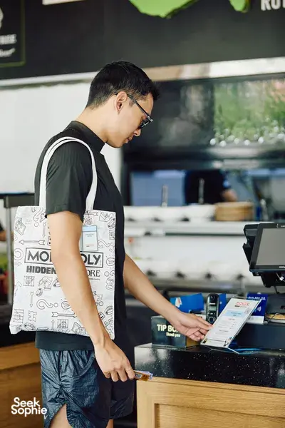 Solo player in a real-life Monopoly game at a Singapore River counter, checking the booklet and grabbing game items—Singapore hidden gems