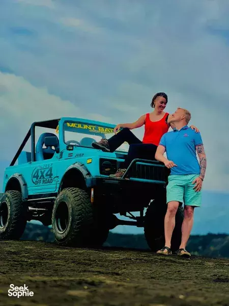 Couple pose beside a turquoise 4x4 jeep against blue mountains in Bali, jeep adventure on Mt Batur