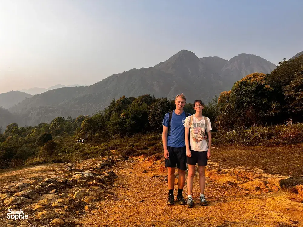 Two travellers walk along a sunlit track through woodland in Sapa, with rugged peaks towering in the distance