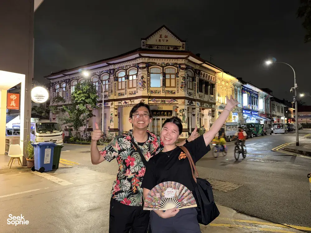 Two people posing on a night street in Geylang, Singapore, holding a folding fan in front of a heritage shophouse