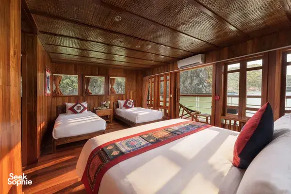 Wooden cabin interior with beds and red cushions, framed by sliding windows to the deck on a boutique cruise in Cat Ba