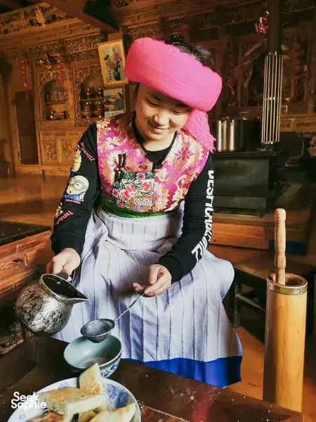 Woman in a pink headscarf pouring tea from a small kettle at a traditional Tibetan home visit in Shangri-La