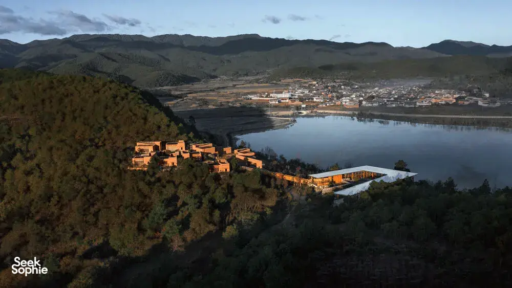 Pemandangan udara Linden Centre eco retreat di Shaxi, Dali, dengan bangunan beratap oranye di tepi danau tenang dan dikelilingi pegunungan