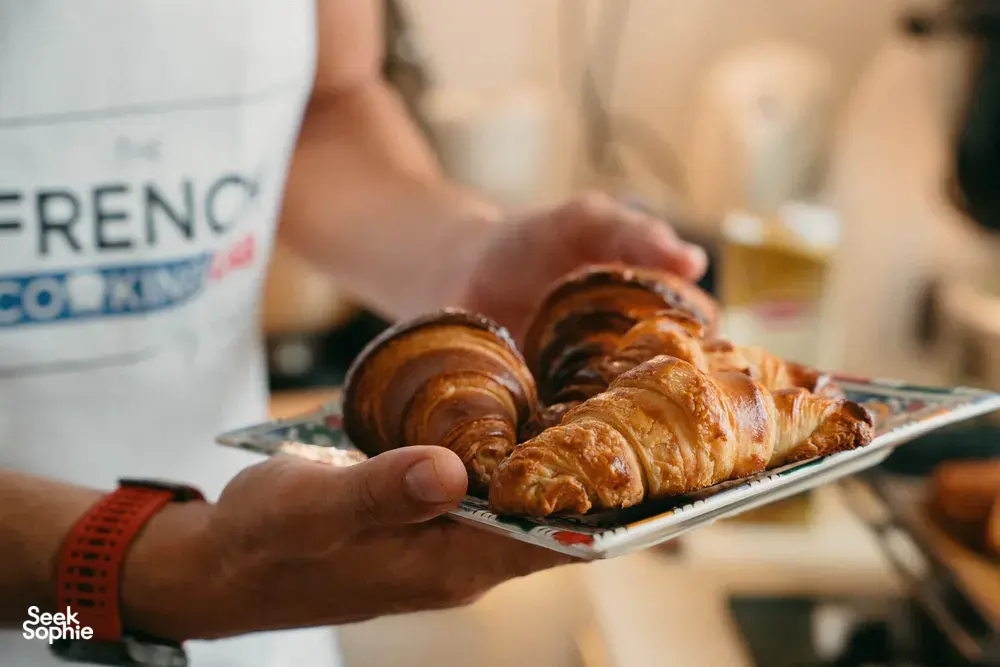 French chef in a French Cooking Lab apron presenting freshly baked golden croissants on a tray in Singapore