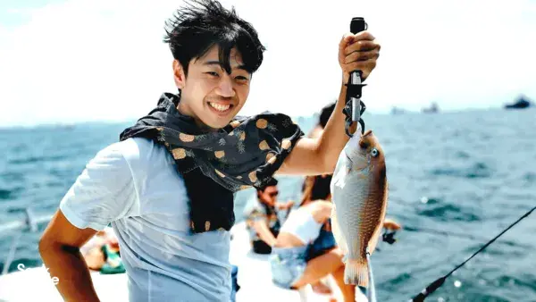 Smiling angler on a Singapore fishing yacht holding a freshly caught fish, with other lines dropping in the background