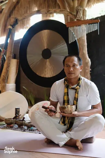 Pranasanti Yoga practitioner seated with a singing bowl in a thatched meditation room in Bali, with a gong behind him in Ubud