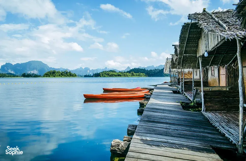 Wooden rafthouse jetty with moored orange kayaks on Cheow Lan Lake, viewed from the water at Khao Sok Riverside Cottages