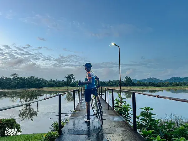Cyclist riding a bike on a bridge over a calm lake near Khao Lak, with lush trees and hills under a blue sky