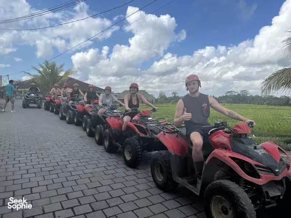 Line of riders on red ATV quad bikes on a Bali cobblestone road, with grassy fields and cloud-filled sky
