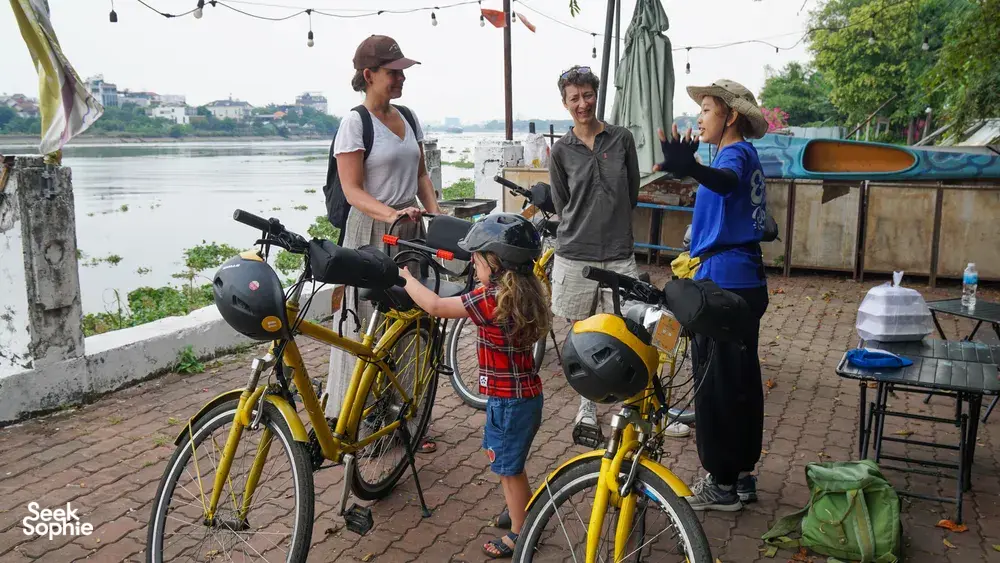 Travellers on yellow bikes pause by the riverside on a Mekong Island trip from Ho Chi Minh City, with a boat behind