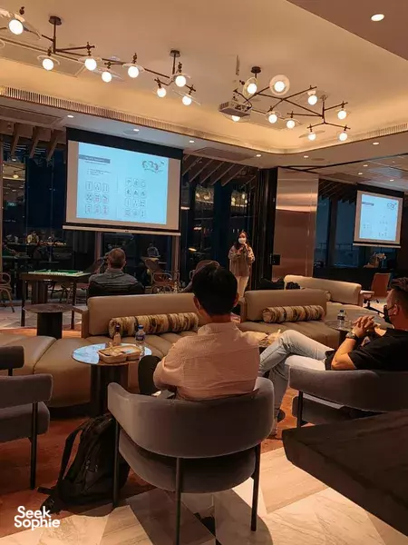 Presenter at a Singapore lounge-style room briefing a team mahjong session, with seated guests facing a screen — workshops in Singapore