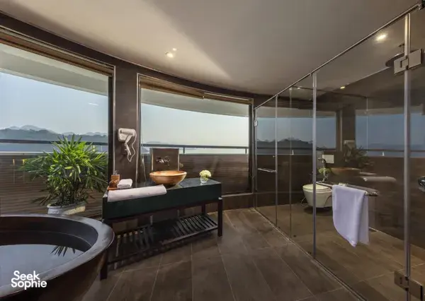 Glass-walled shower and bathtub in an Oasis Bay Party Cruise suite, with windows overlooking Halong Bay scenery