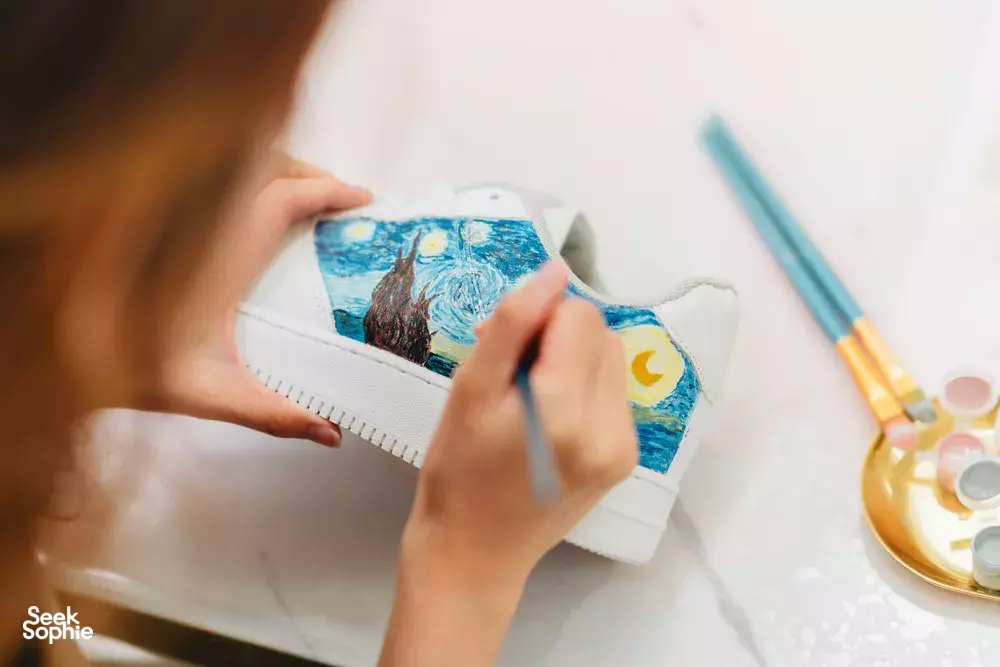 Hands sketching a bright ocean scene on a small canvas notebook at a creative table, with pencils and paint for workshops in Singapore