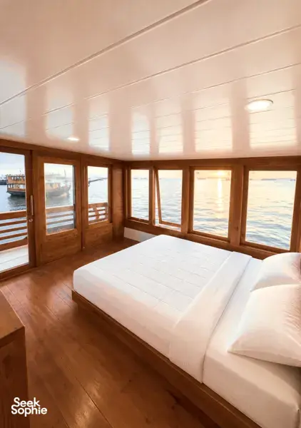 White-mattress bed in a wooden phinisi cabin with floor-to-ceiling windows, part of a 3D2N shared Komodo cruise from Labuan Bajo