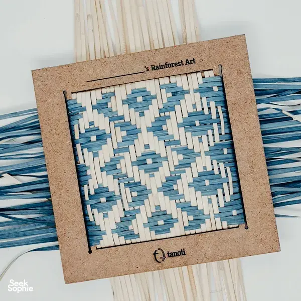 Framed woven artwork on cork at Tanoti Crafts in Kuching, displaying a blue-and-white rainforest-style weave pattern