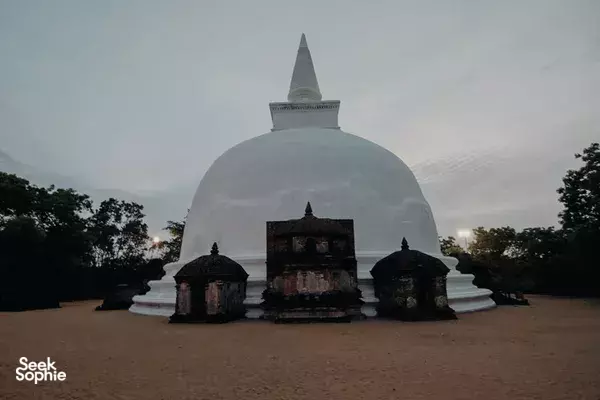 White domed stupa and smaller shrines in a grassy courtyard at dusk, set among trees in Polonnaruwa