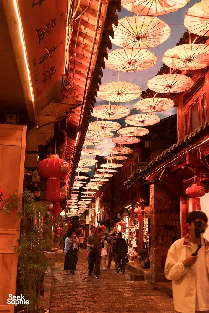 Lantern-covered alley in Lijiang Old Town, warm red light beneath hanging parasols as visitors stroll through the narrow lane